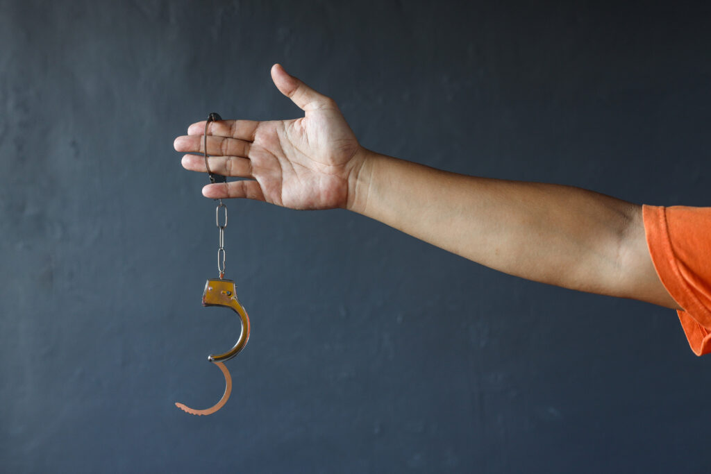 Photo of a man’s outstretched arm. His wrist is in one cuff of a set of handcuffs, the other cuff dangles below, unlocked.