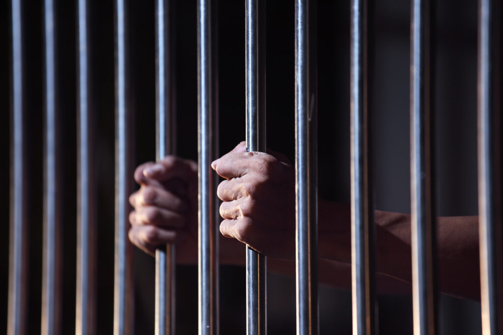 Photo of a man’s hands clutching the bars of a jail cell.