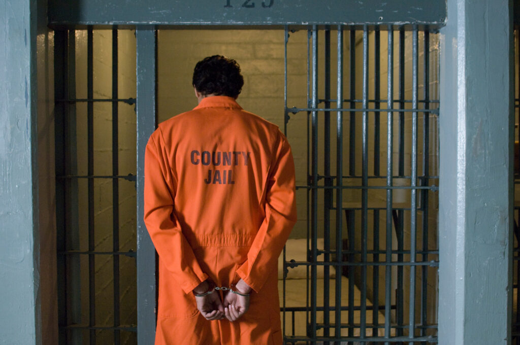 View from behind of a man entering a jail cell. He wears an orange jumpsuit with the words “COUNTY JAIL” printed on the back.