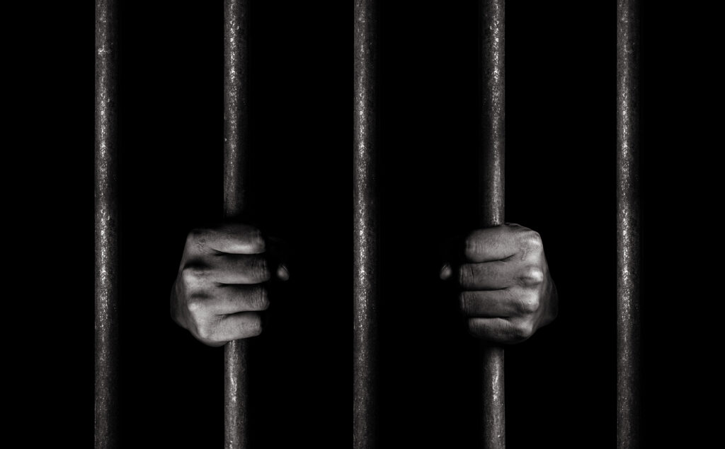 Image of a dark jail cell, the only things visible are the bars and a pair of hands gripping two of the bars.